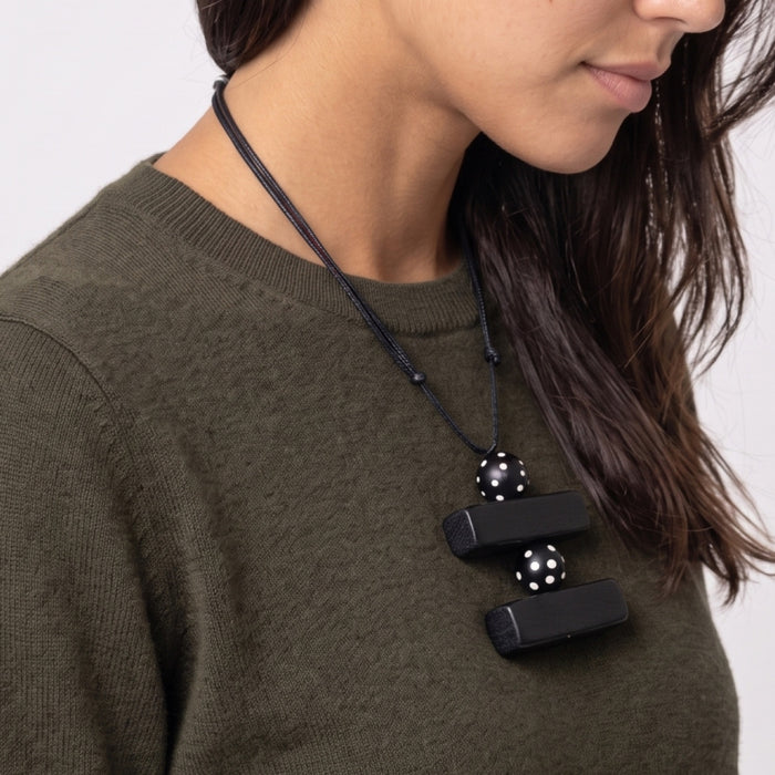 A close-up of a woman wearing a black cord necklace with two black rectangular beads, each detailed with white polka dots, over an olive green knit sweater.