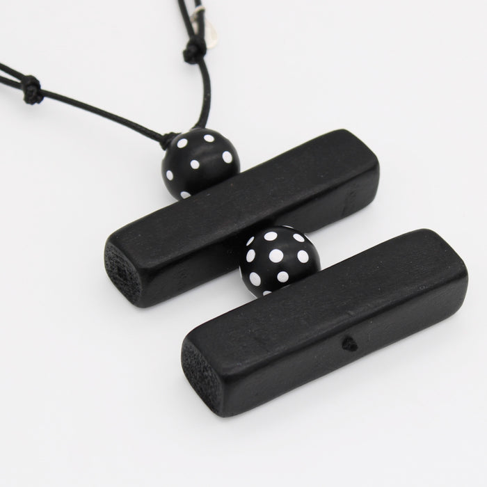 Black wooden pendant necklace with two rectangular bars connected by a black cord and two black beads with white polka dots, shown on a white background.