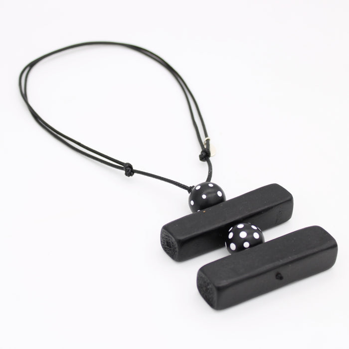 Black wooden pendant necklace with two rectangular bars connected by a black cord and accented with two black beads featuring white polka dots, shown on a plain white background.