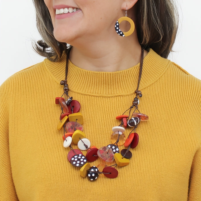 A close-up of a woman wearing a colorful mushroom-themed statement necklace and matching drop earrings. The necklace features various wooden buttons and discs in red, yellow, white, and black with polka dots, strung on a dark brown cord.