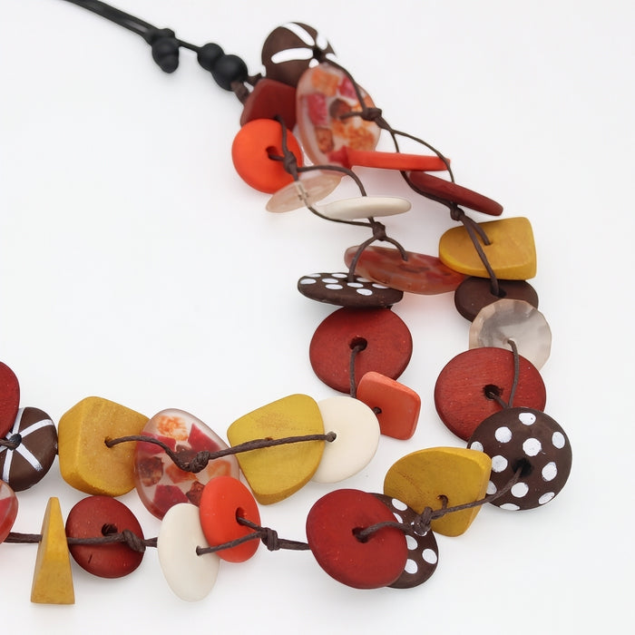 A multi-strand artisan necklace featuring organic-shaped wooden beads in red, yellow, white, and brown, some with polka dots or marbled patterns, threaded on dark brown cords against a white background.