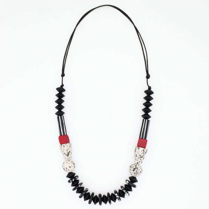 A beaded necklace featuring black faceted beads, black and white striped sections, textured silver-toned details, and small red accent beads, suspended on a black cord against a plain white background.