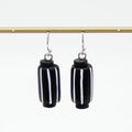 A pair of black cylindrical drop earrings with vertical white stripes, hanging from silver-toned hooks from a gold horizontal bar against a white background.