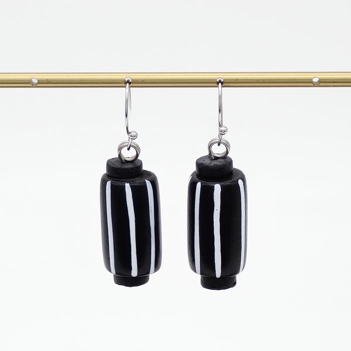 A pair of black cylindrical drop earrings with vertical white stripes, hanging from silver-toned hooks from a gold horizontal bar against a white background.