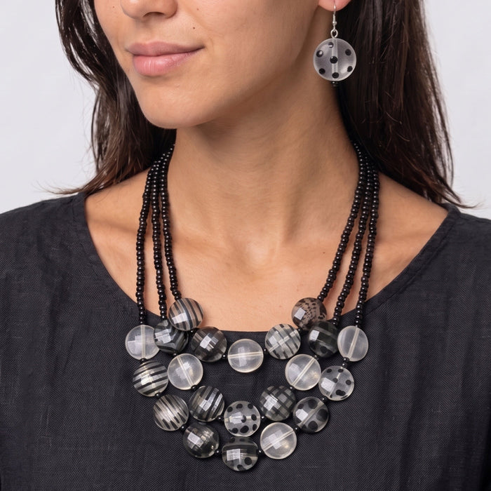 Woman wearing A multi-strand statement necklace featuring black beaded cords and a tiered arrangement of round beads in black, white, and gray patterns, including stripes, polka dots, and marbled textures, on a plain white background.