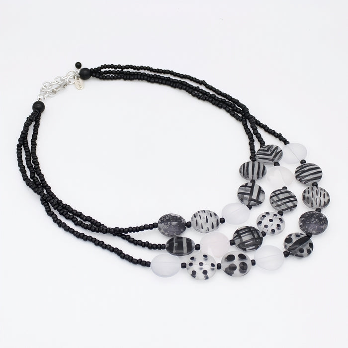A multi-strand statement necklace featuring black beaded strands, translucent white beads, and patterned black-and-white disc beads with stripes, dots, and marbled textures, finished with a silver-tone clasp.
