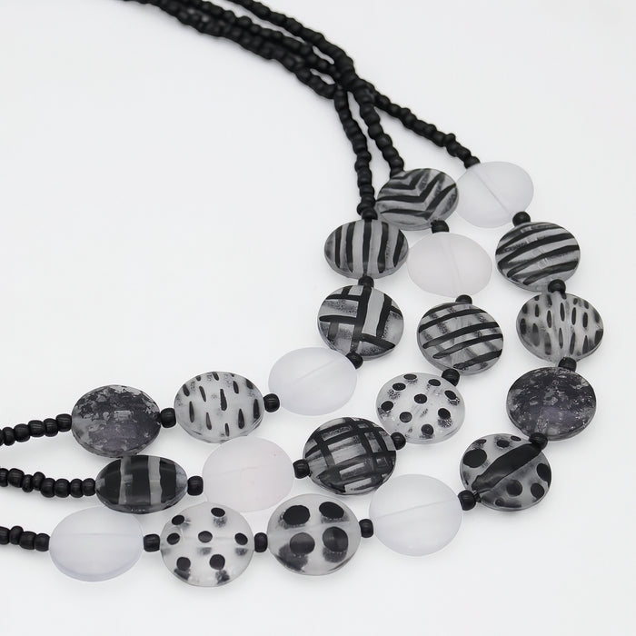 A multi-strand beaded necklace featuring black and white patterned round beads, translucent white beads, and black cord, displayed on a clean white background.