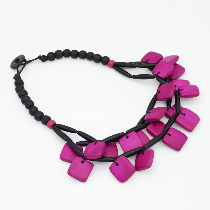 A statement necklace featuring bright magenta-pink square beads and black cylindrical links, displayed on a clean white background.