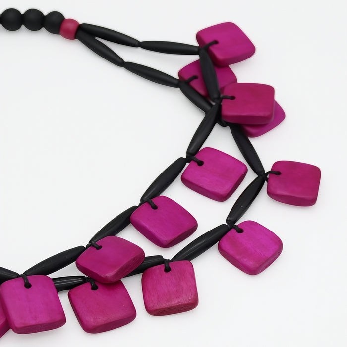 A close-up of a modern statement necklace featuring bright magenta-pink square wooden beads strung on a black cord, set against a plain white background.