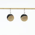 A pair of modern dangle earrings with a split black and golf design, hanging from silver hooks on a gold horizontal bar against a white background.