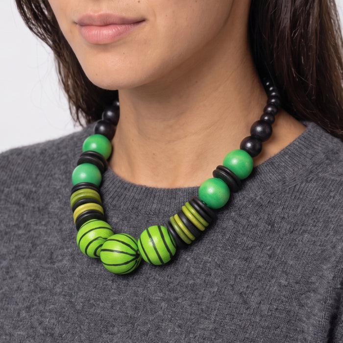 A close-up of a woman wearing a chunky statement necklace made of green and black wooden beads, including striped and solid green beads, paired with a gray knit sweater.