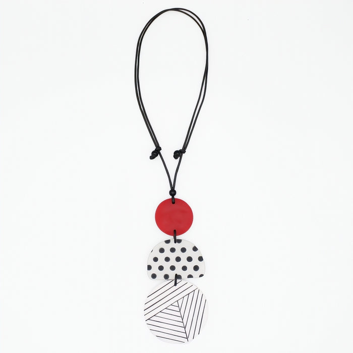 A modern multi-piece pendant necklace featuring a black cord, a solid red disc, a white disc with black polka dots, and a white disc with black diagonal lines, shown on a plain white background.
