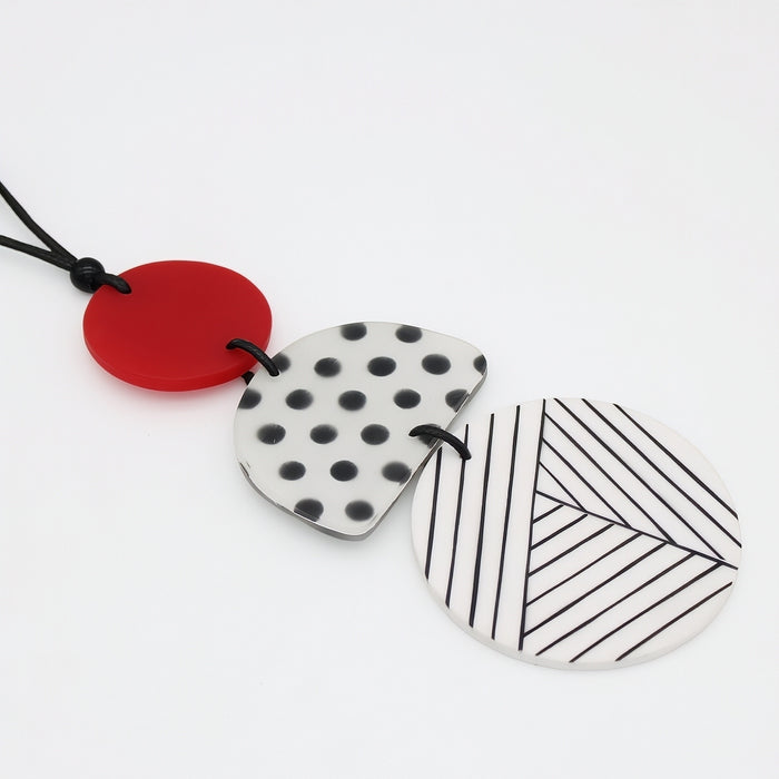 Three circular pendant necklaces with black cords, featuring a solid red pendant, a white pendant with black polka dots, and a white pendant with black diagonal stripes, arranged on a clean white background.