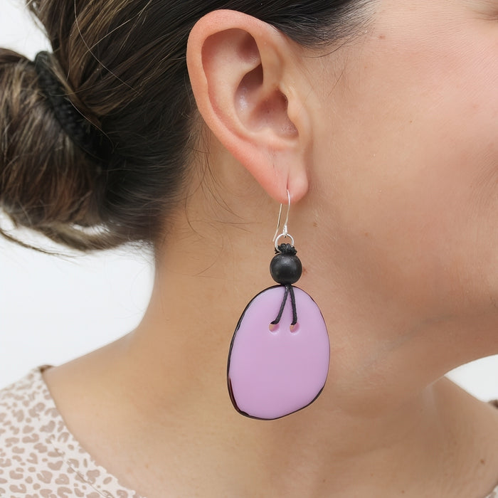 A close-up of a person's ear wearing a dangling earring. The earring has a silver fishhook wire, a small black bead, and an irregular oval drop made from light pink material with black stitched details.