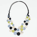 A multi-strand statement necklace featuring circular wooden discs in black, light blue, olive green, and cream with abstract line patterns, suspended on a black cord against a white background.