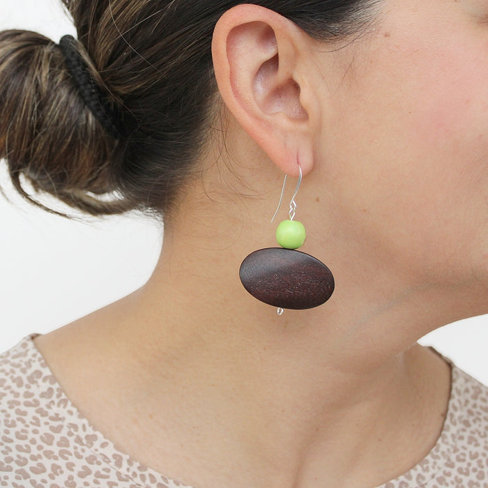 A close-up of a person's ear wearing a dangling earring with a small light green bead and a larger dark brown oval drop, against a plain light background.