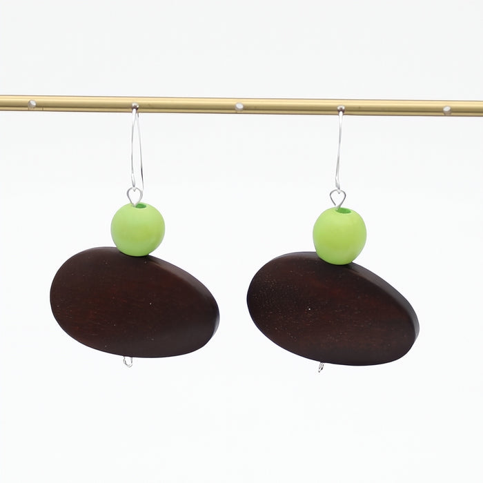 A pair of dangle earrings with dark brown oval stone pendants, a bright lime green round bead, and silver fishhook wires, hanging from a gold-colored display rod against a white background.
