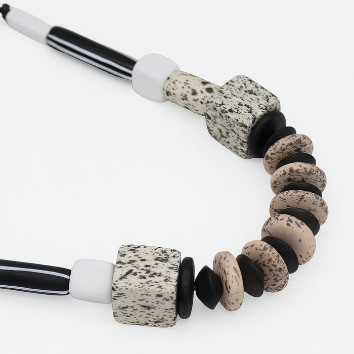 A close-up of a beaded necklace featuring speckled stone beads in beige, black, and white, with striped black and white cord ends, shot against a clean white background.