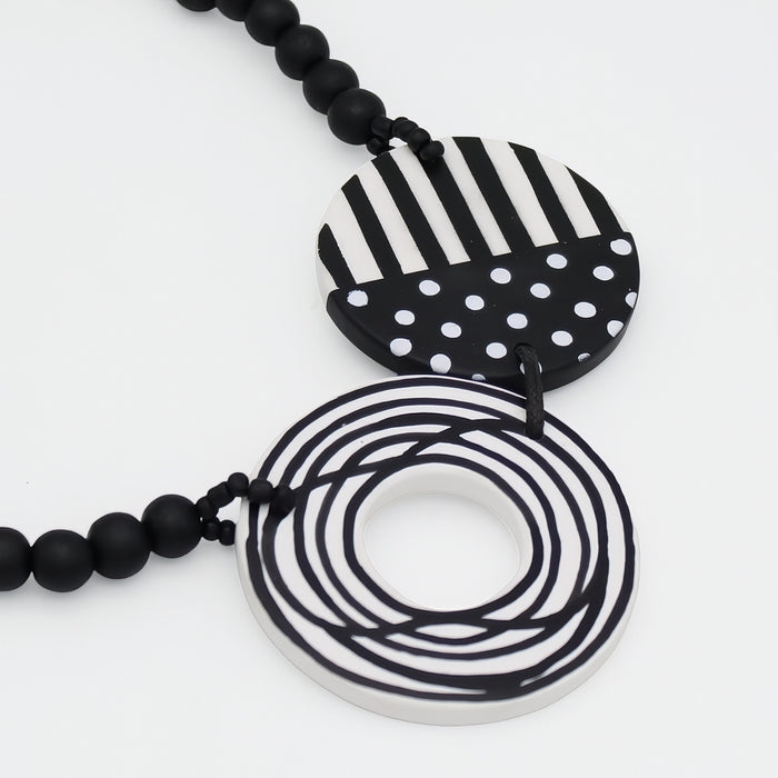 Black and white geometric pendant necklace featuring a striped and polka-dot circular pendant and a coiled ring pendant, suspended over a black beaded chain on a white background.