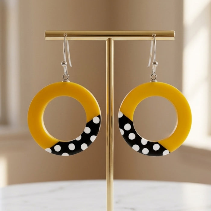 A pair of yellow circular drop earrings with a black and white polka dot section at the bottom, hanging from silver hooks on a gold T-stand against a softly blurred beige background.