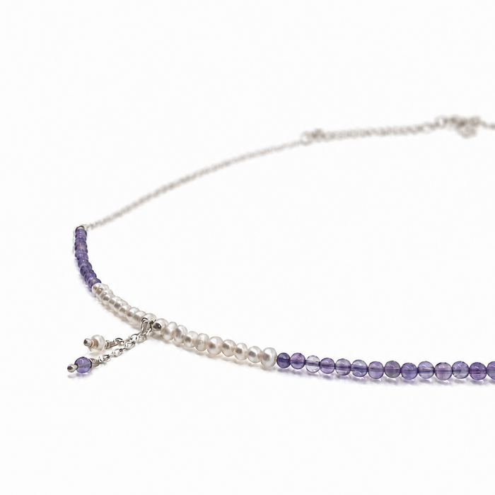 A delicate silver-toned necklace featuring a strand of white pearls and a strand of purple beads, with a small dangling charm, shown on a plain white background.