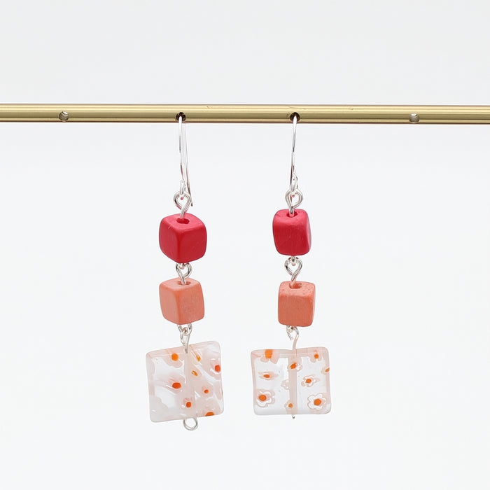 A pair of dangling earrings hanging from a gold rod against a white background. Each earring has a silver fishhook wire, a small red cube bead, a peach-orange cube bead, and a square white glass tile with orange speckles.