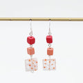 A pair of dangling earrings hanging from a gold rod against a white background. Each earring has a silver fishhook wire, a small red cube bead, a peach-orange cube bead, and a square white glass tile with orange speckles.