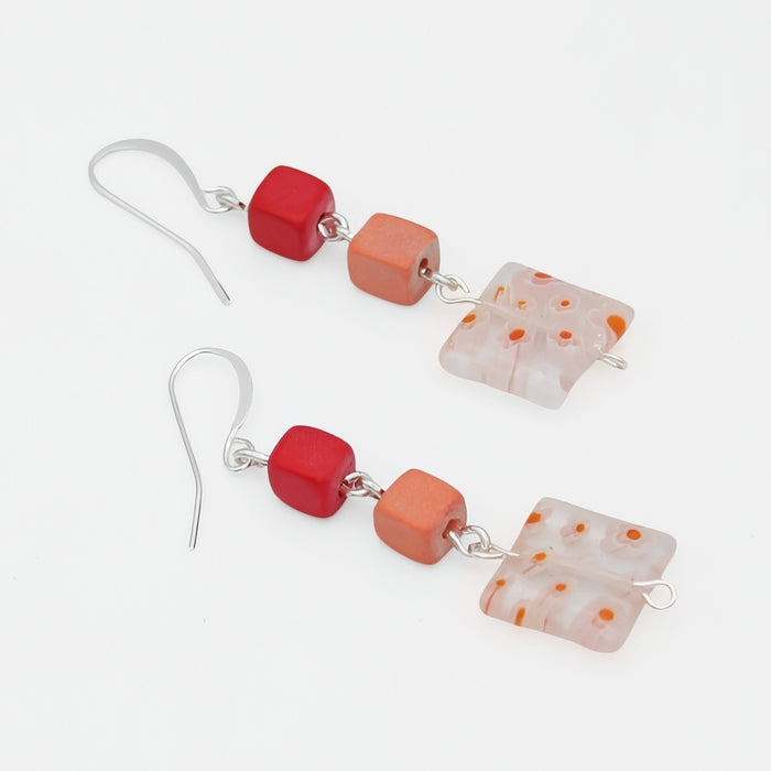 A pair of handmade dangle earrings featuring silver fishhook wires, a glossy red cube bead, a matte orange cube bead, and a translucent white square glass bead with orange speckles, displayed on a clean white background.