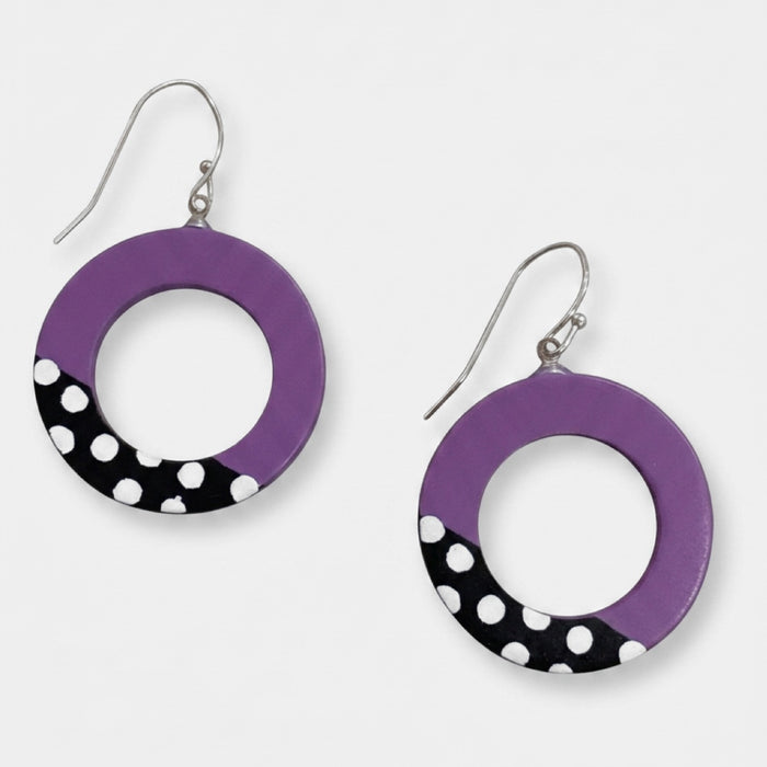 A pair of RESIN hoop earrings with a purple top half and a black bottom half decorated with white polka dots, hanging from silver fishhook wires on a light gray background.
