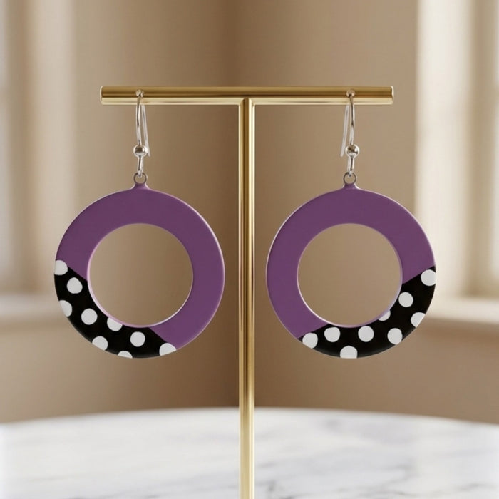 Purple and black hoop earrings with white polka dots, hanging from a gold T-stand against a soft beige background.