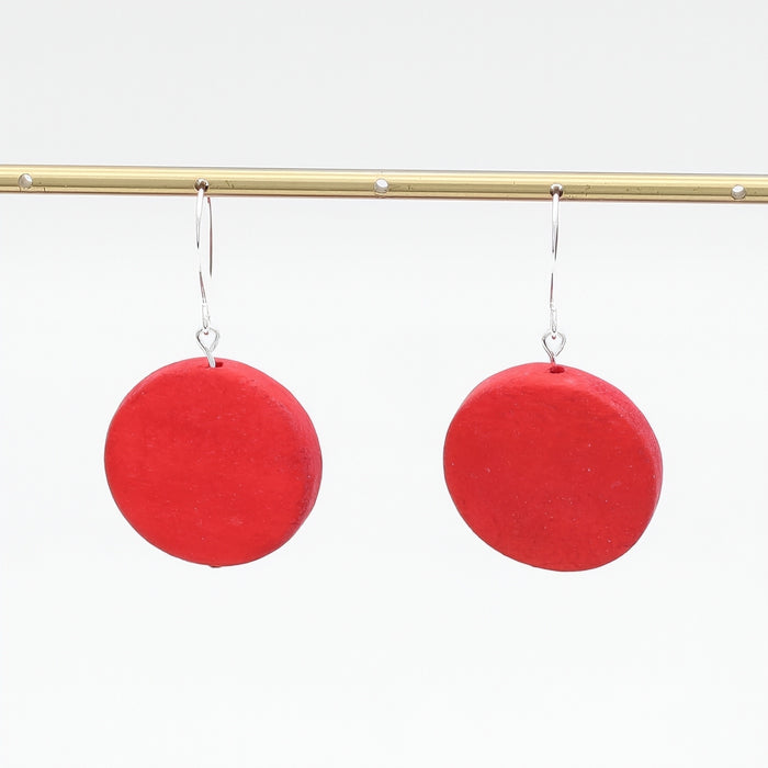 A pair of dangle earrings featuring round, matte red discs suspended from thin silver fishhook wires, set against a clean white background.