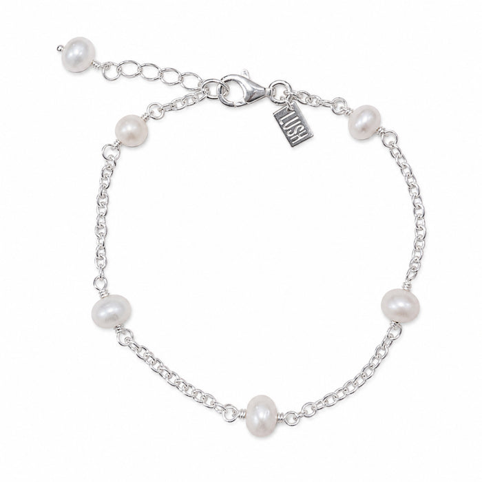 A silver chain bracelet with six white freshwater pearls spaced evenly along the chain, featuring a lobster clasp, extender chain, and a small engraved brand tag that reads 'LUSH'.