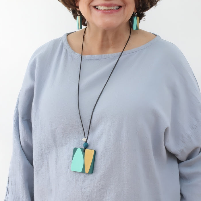 Woman wearing a light blue shirt with a long necklace featuring a colorful pendant against a white background