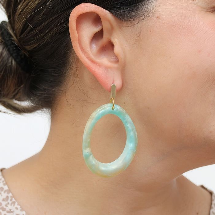 A close-up of a person's ear wearing a large, oval drop earring made of marbled light blue and green resin with a small gold-tone hook.