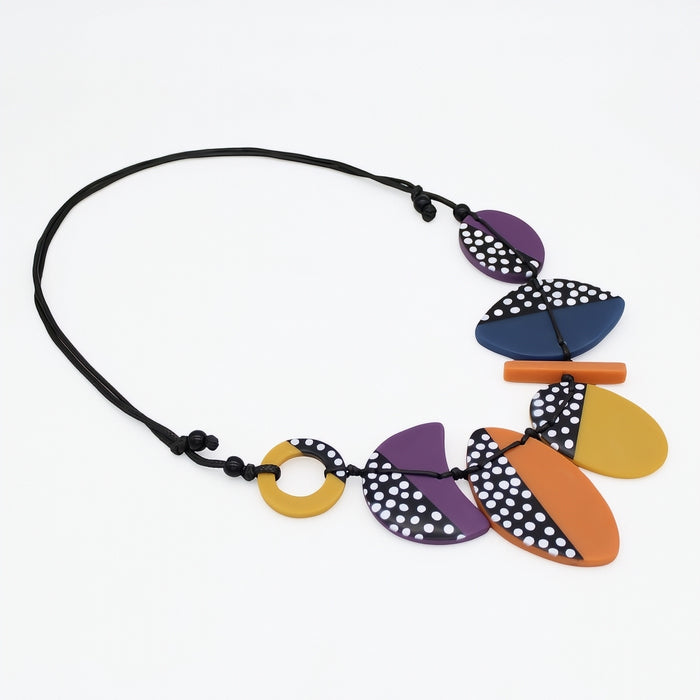 A modern multi-piece necklace featuring abstract organic shapes in purple, orange, mustard yellow, and navy blue, some with black and white polka dots, suspended on a black cord against a white background.