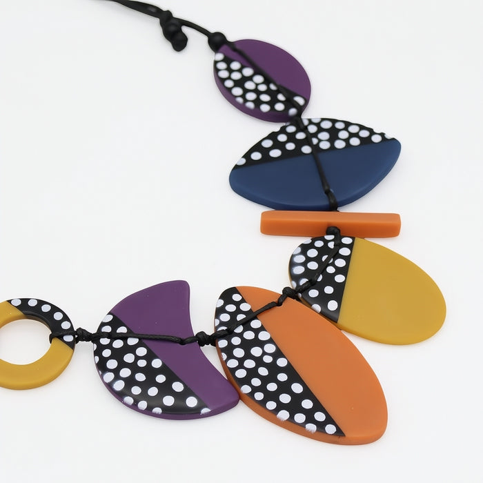 A colorful geometric pendant necklace on a black cord, featuring semi-circular and teardrop-shaped beads in purple, navy blue, orange, and mustard yellow, with some beads decorated in a black and white polka dot pattern.