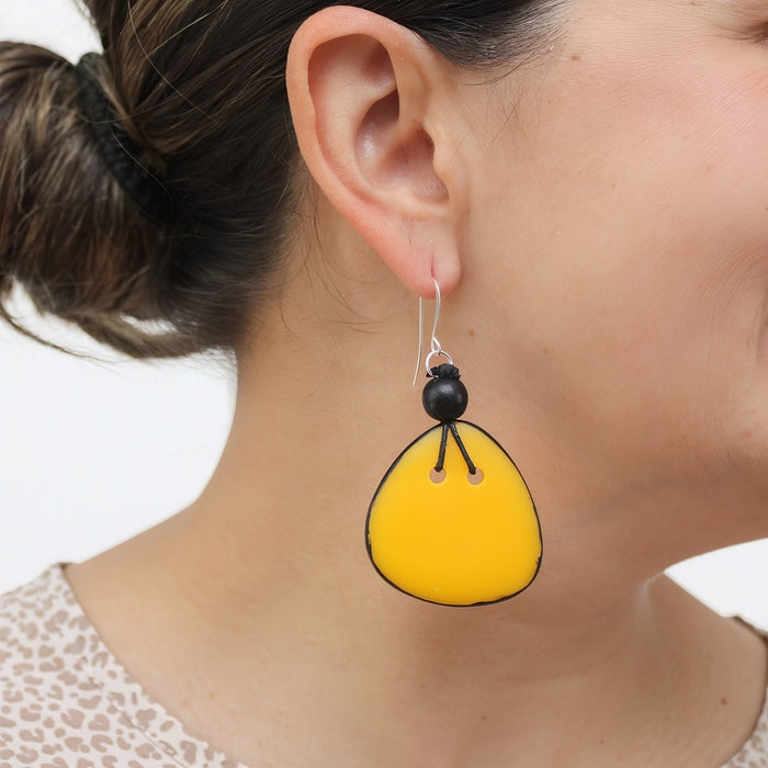 A close-up of a woman's ear wearing a dangling earring. The earring has a bright yellow teardrop-shaped drop with black hand-painted outlines and a small black bead above it on a silver hook.