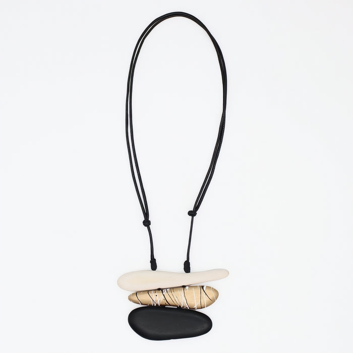 A minimalist necklace featuring three stacked natural stones in cream, tan, and black, suspended on a black adjustable cord against a plain white background.