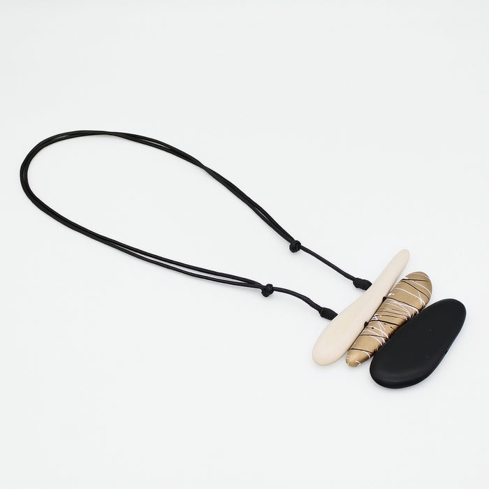 A modern pendant necklace featuring a black adjustable cord, a flat cream-colored stone, a textured gold and beige wrapped stone, and a smooth black stone, displayed on a clean white background.