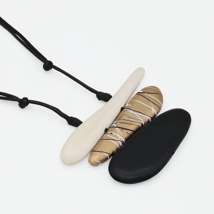 Three flat oval pendant necklaces on black cords, displayed on a clean white background. The necklaces feature matte black, matte cream, and a textured tan-and-white patterned pendant.