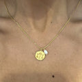 A gold necklace with a Libra zodiac sign pendant and a dainty pearl suspended from the chain, worn around the neck.